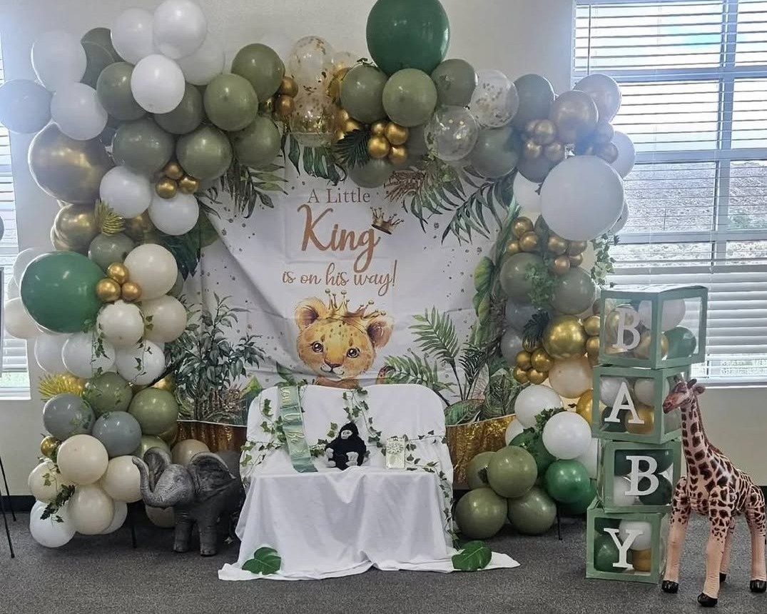 A sage green and gold balloon garland framing a jungle-themed baby shower backdrop reading A Little King is on His Way