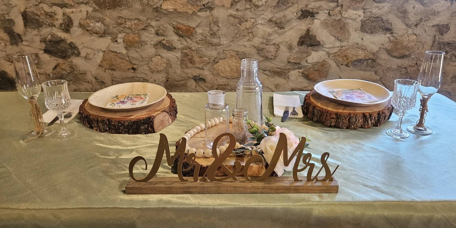 A rustic wedding place setting with a wood charger, sage tablecloth, and a gold Mr. and Mrs. sign against a stone wall