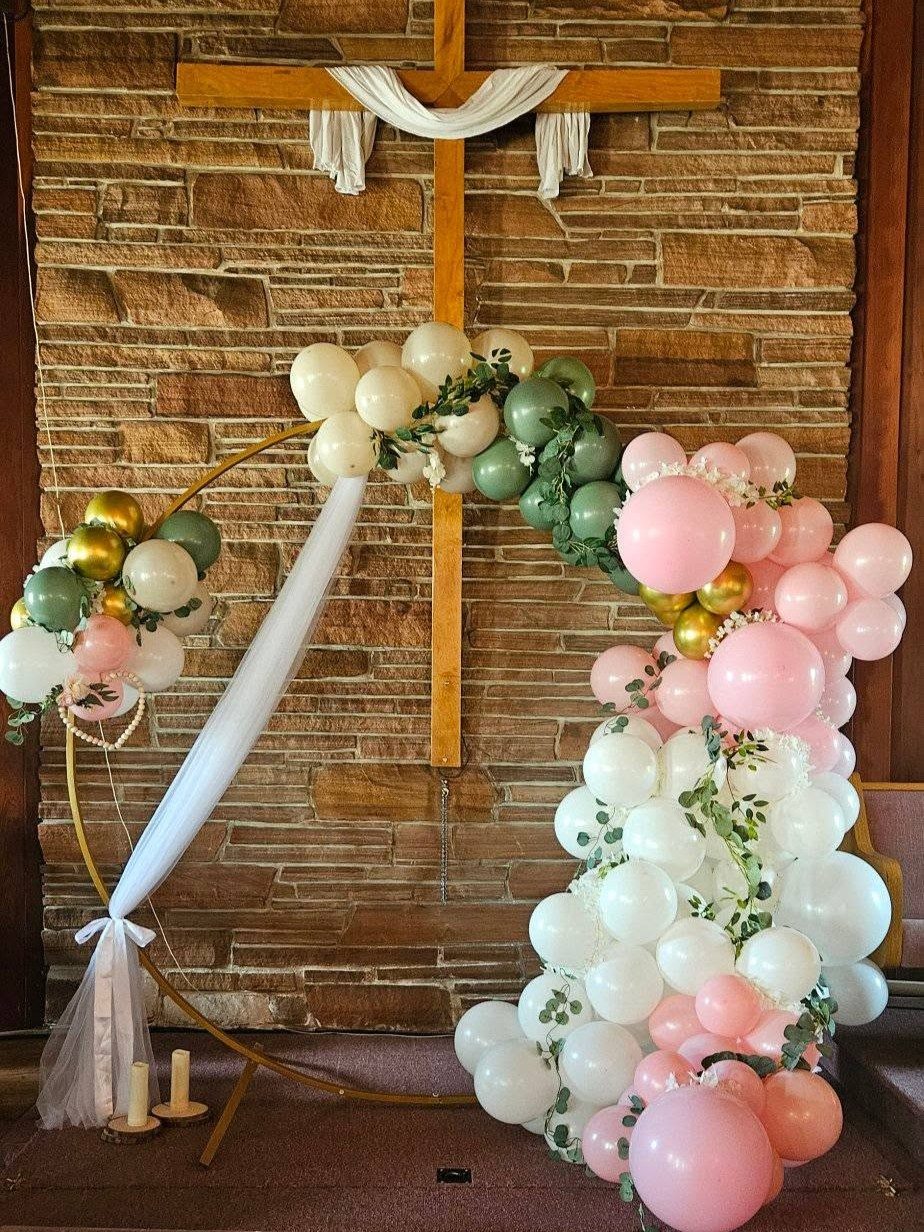 A sage-green, gold, and white organic balloon garland arcing over a wooden cross at a stone-walled venue