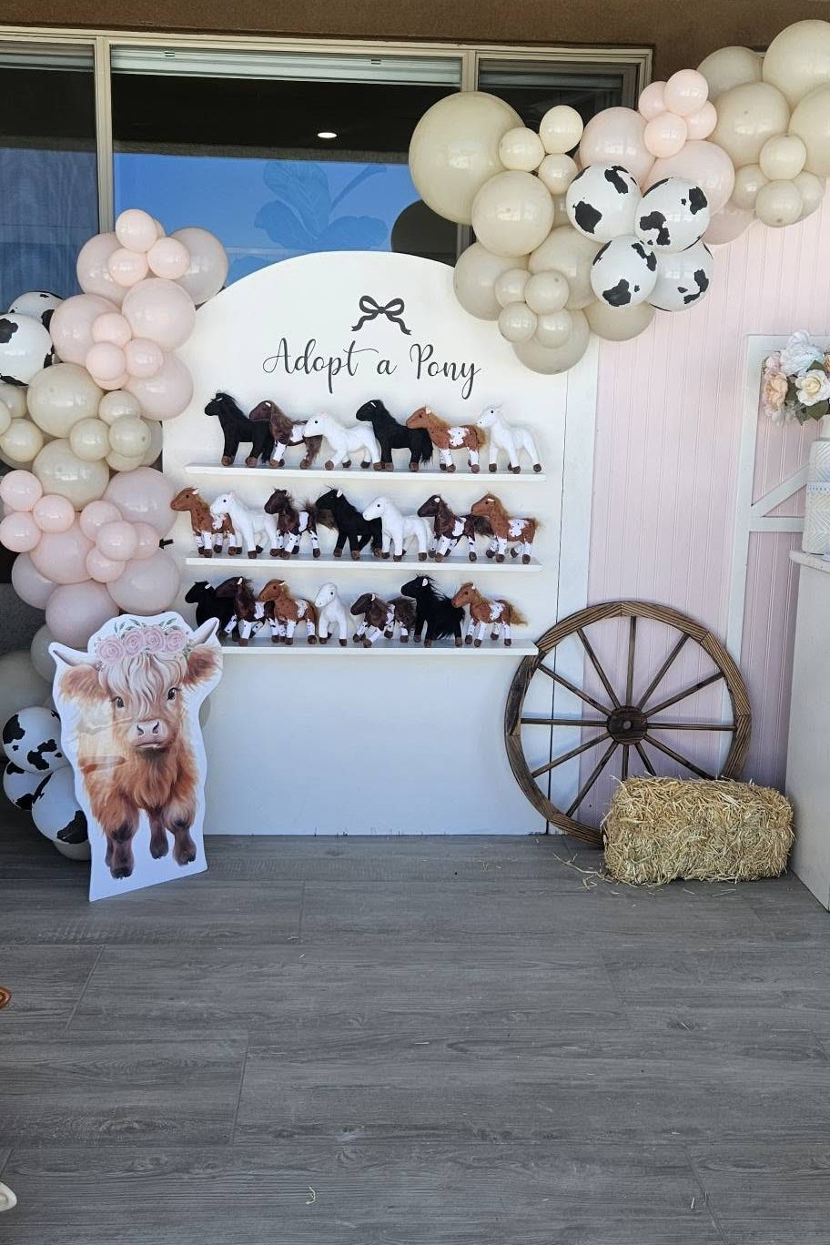 An Adopt a Pony backdrop under a cream, blush, and cow-print balloon garland, with plush ponies on shelves beside a wagon wheel and hay bale