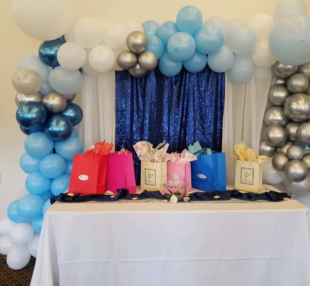 A blue sequin backdrop framed by a navy, ice blue, and chrome balloon garland, above a gift table lined with favour bags