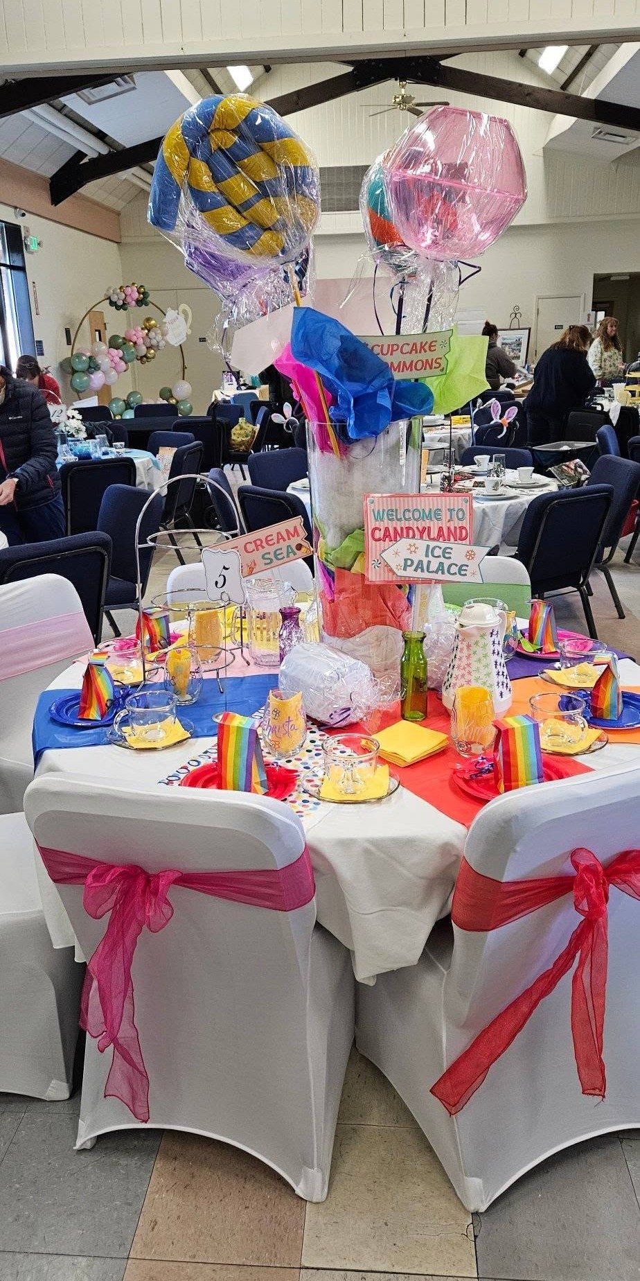 A children's party room with red and blue tables, tied chair covers, balloon centerpieces, and carnival treats