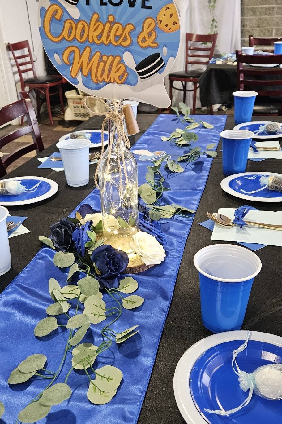 A long baby shower table with a royal blue satin runner over black linen, blue and white plates, eucalyptus, and an I Love Cookies and Milk sign