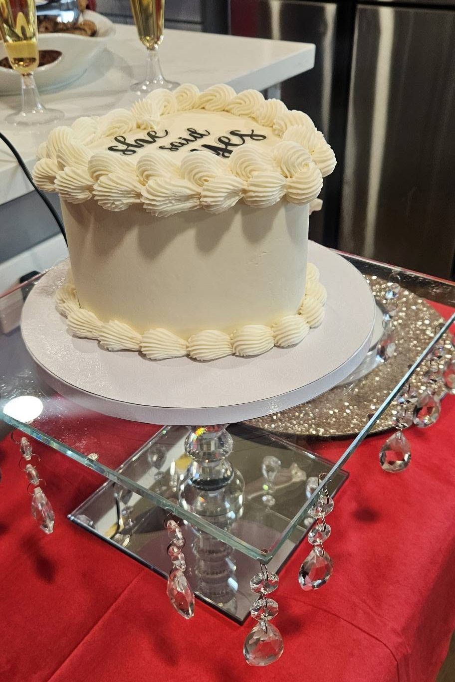 A single-tier white cake lettered She Said Yes on a mirrored crystal riser over red linen, with champagne poured behind it