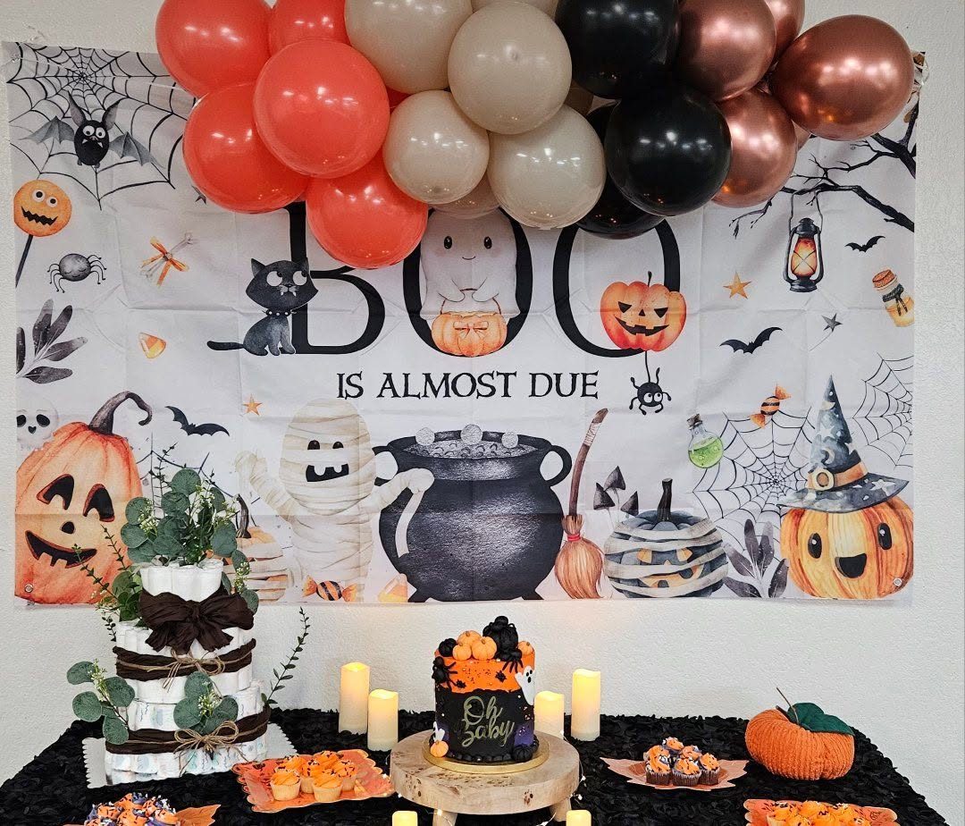 A Halloween baby shower dessert table under a Boo Is Almost Due backdrop, with coral, black, and rose gold balloons, a naked cake, and pumpkins