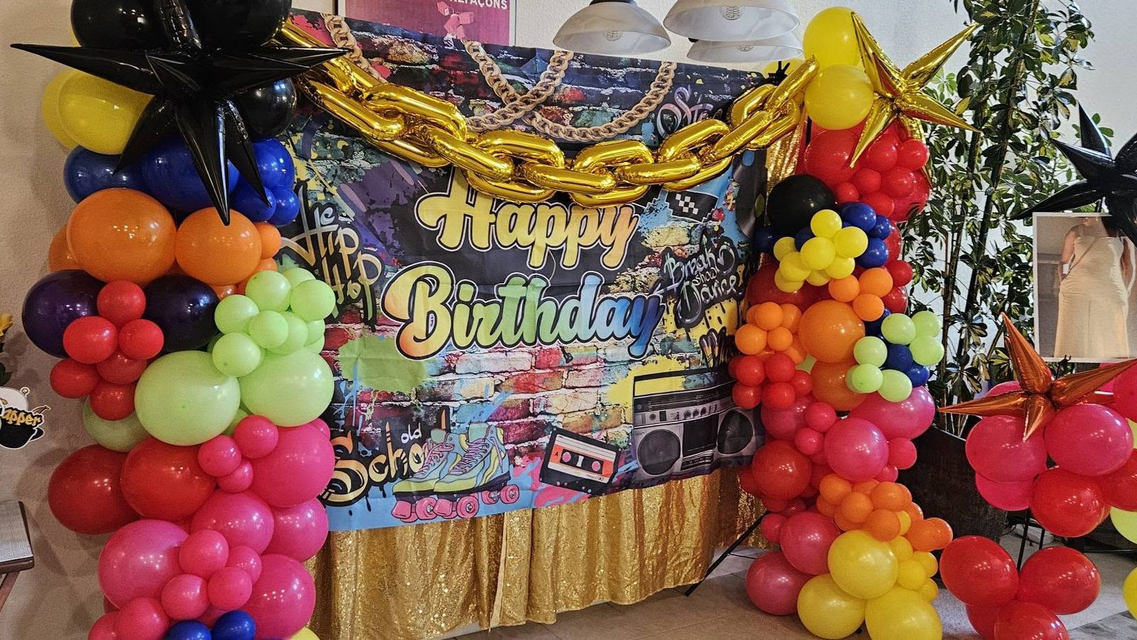 A retro hip-hop birthday backdrop with a boombox and gold chain graphic over a gold sequin skirt, framed by red, blue, and yellow balloon columns