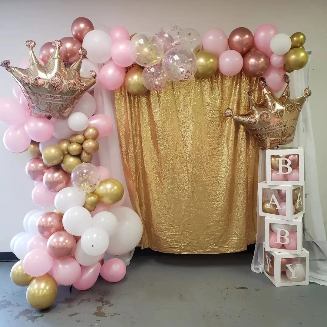 A gold sequin backdrop framed by a blush, white, and rose gold balloon garland with foil crowns, beside clear blocks spelling BABY