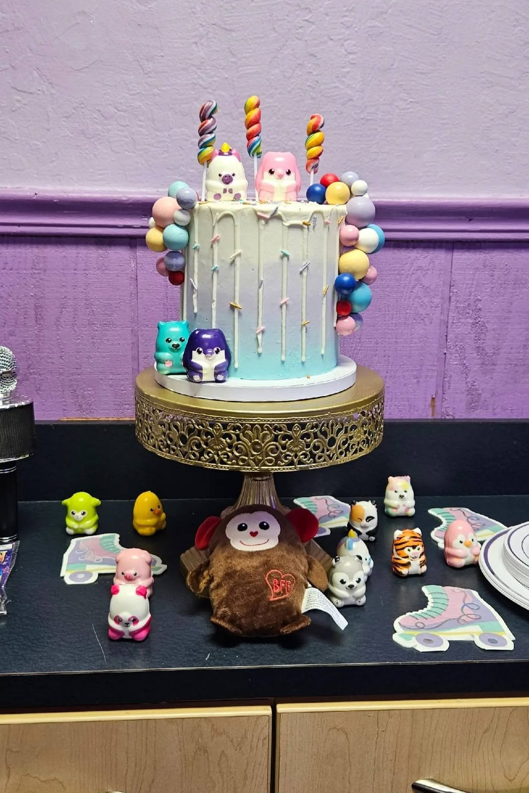 A pastel drip birthday cake topped with candles and small figurines, on a gold stand against a purple wall