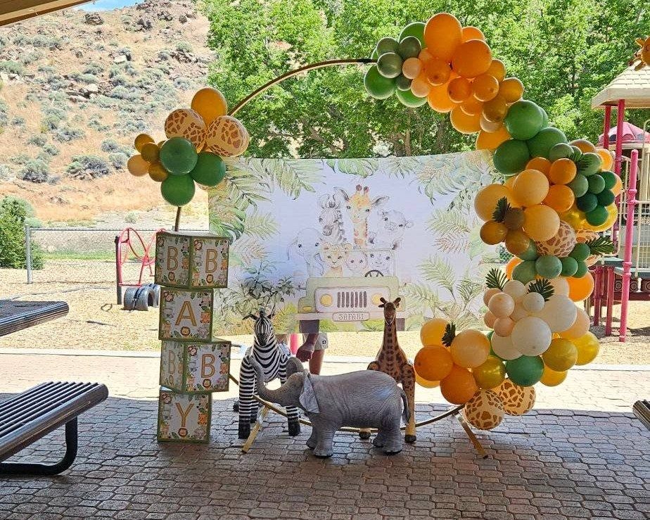 A green, orange, and giraffe-print balloon arch over a safari backdrop with animal figures at an outdoor park