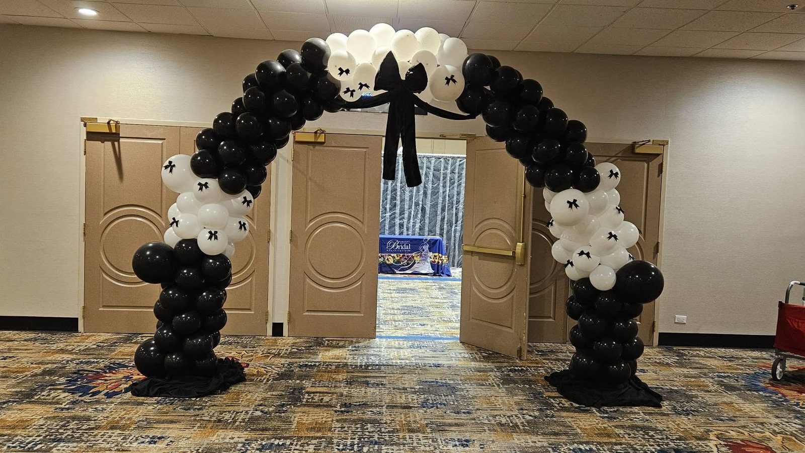 A black and white balloon arch spanning double banquet-room doors with a black satin bow at the crown
