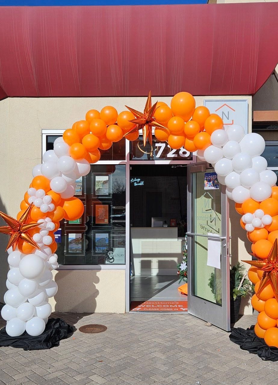 An orange and white balloon arch with copper starburst accents framing the entrance of a real-estate storefront under a red awning