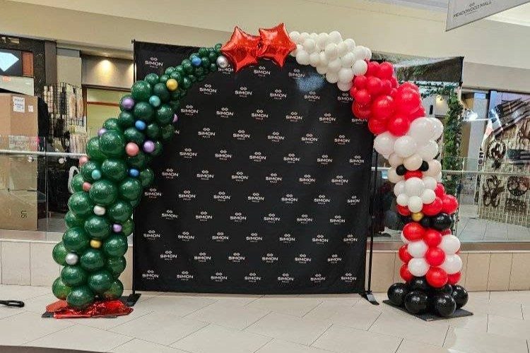A green, red, and white Christmas balloon arch with stars framing a branded step-and-repeat backdrop in a shopping mall