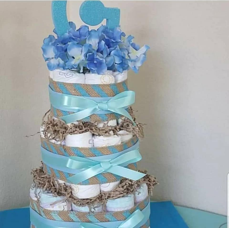 A three-tier diaper cake banded in blue ribbon and burlap with blue hydrangea and a foil monogram topper