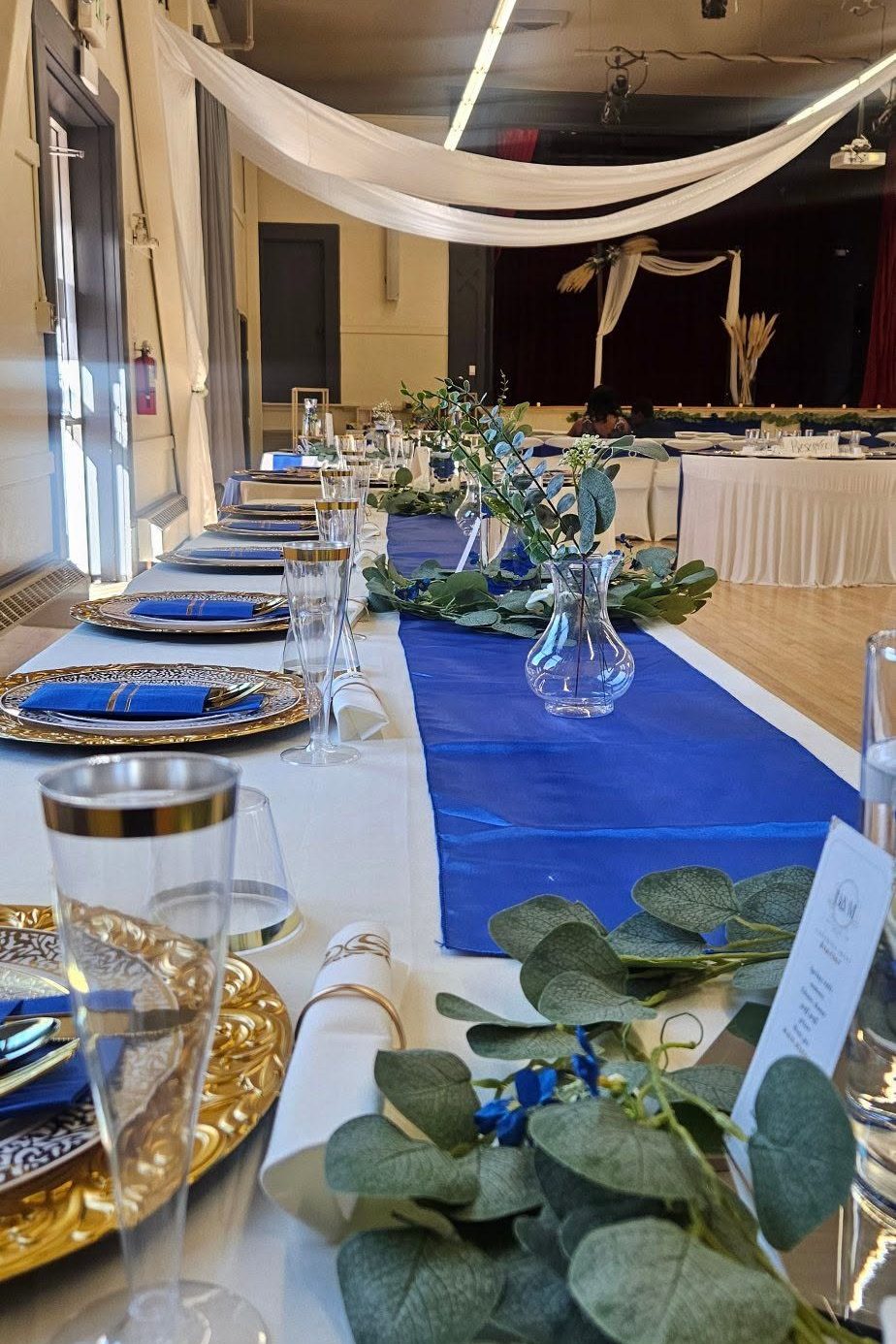 A long table with a royal blue satin runner, gold charger plates, blue napkins, and eucalyptus down the centre