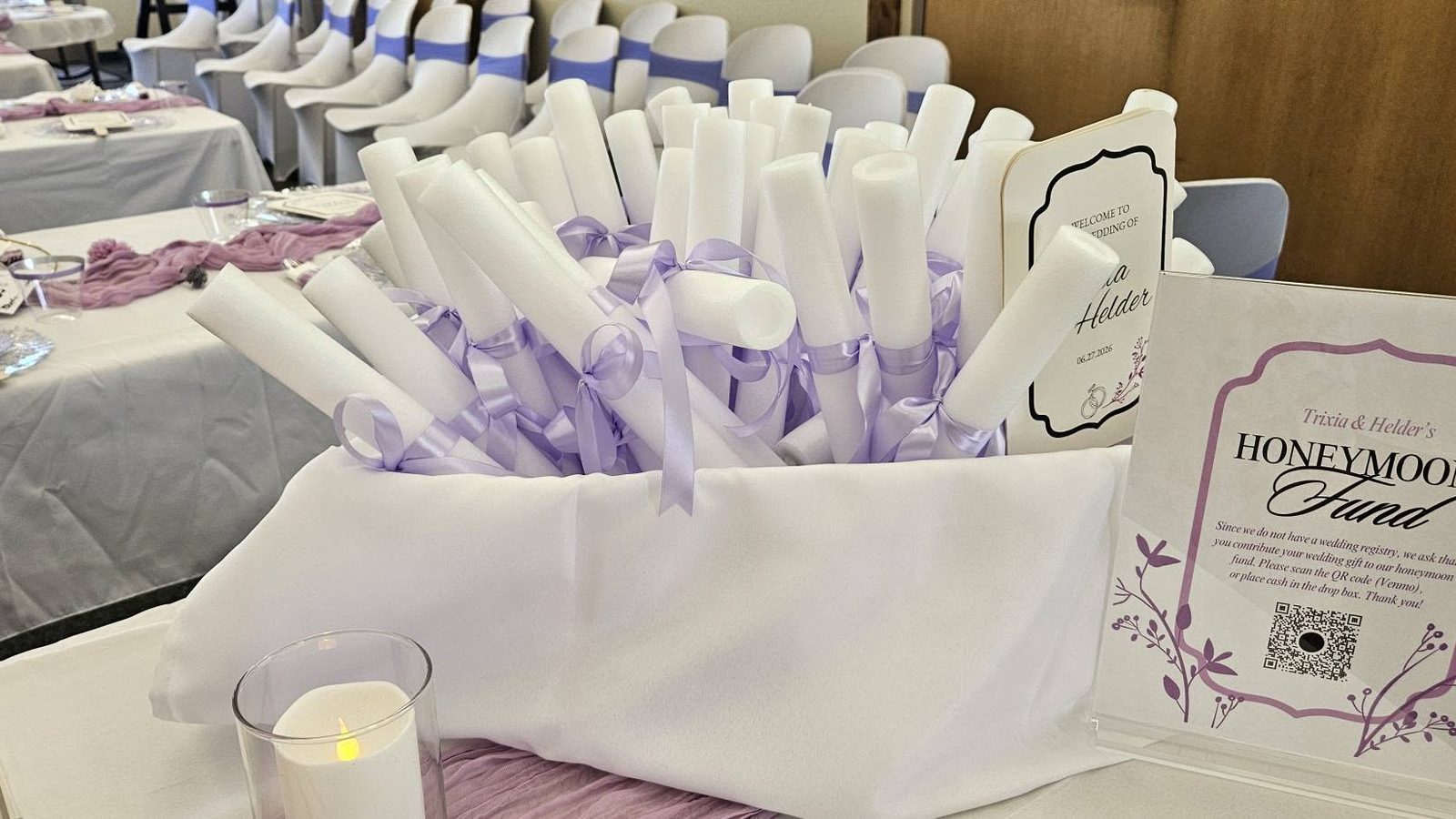 A favour table of rolled lavender takeaways in a white basket beside a framed honeymoon fund sign