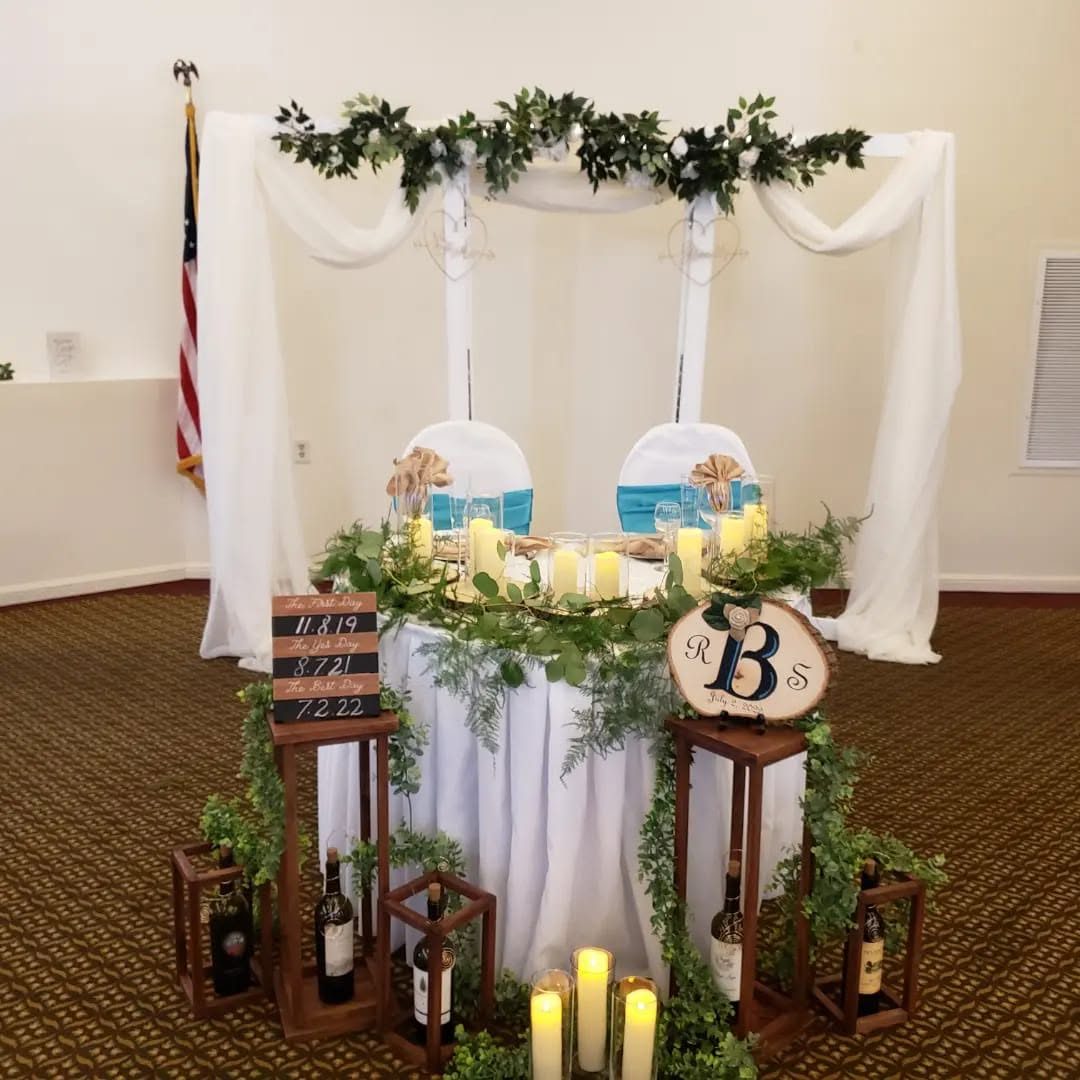 A sweetheart table under white draping with greenery garland, wooden lanterns, pillar candles, and a monogram sign