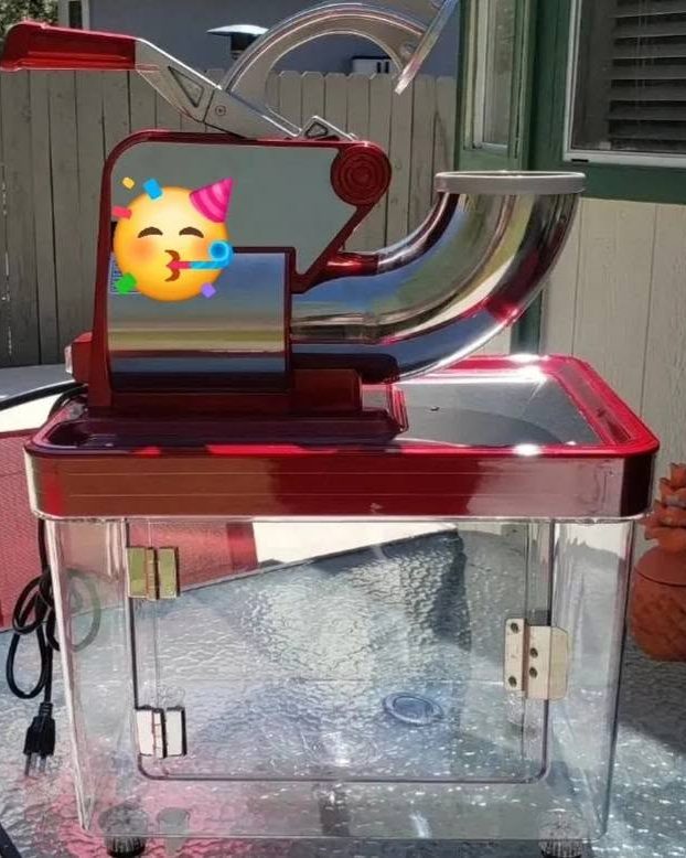 A red and chrome snow cone machine with a clear ice cabinet, set up outdoors