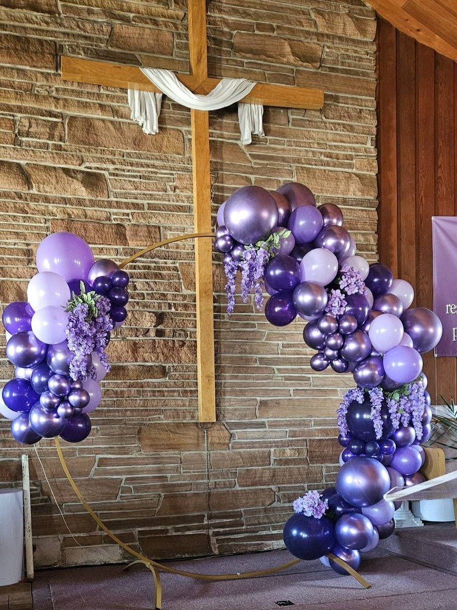 A purple and lavender organic balloon garland with wisteria accents arcing over a wooden cross at a stone-walled venue
