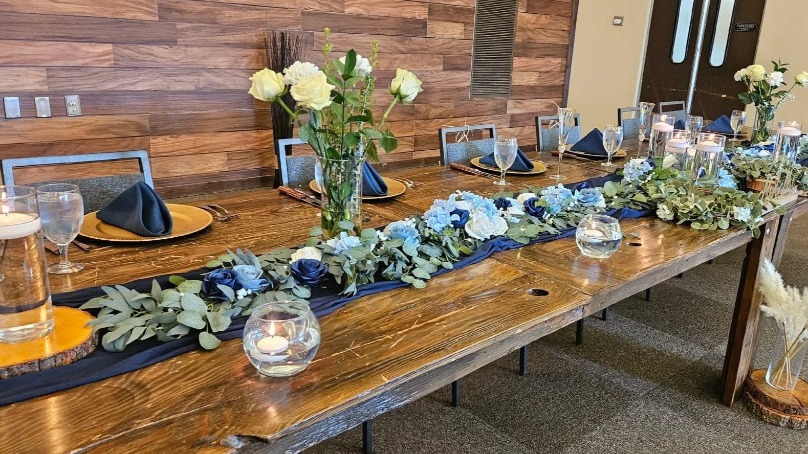 A long wooden bridal table dressed with a navy and blue floral garland runner, candles, and gold-rimmed place settings