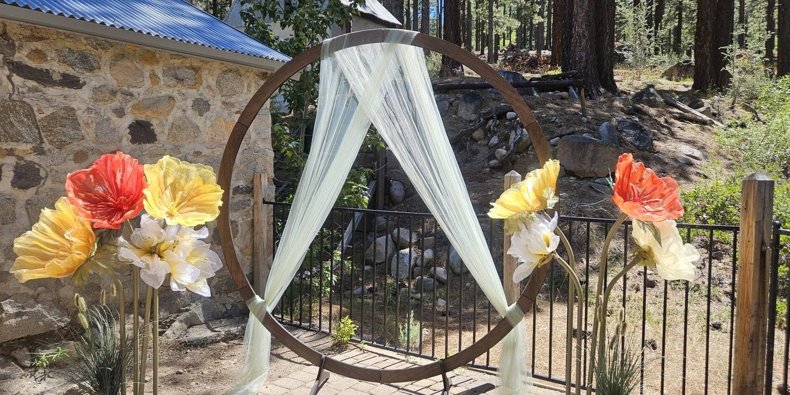 A wooden circular ceremony arch with sheer draping and oversized poppy flowers, set against a stone building and pine forest